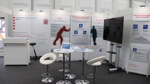 Stand des LfV Hessen beim Hessentag in Bad Hersfeld 2019. Vor einer Messebauwand mit Grafiken stehen ein Tresen und ein Stehtisch mit Barhockern.