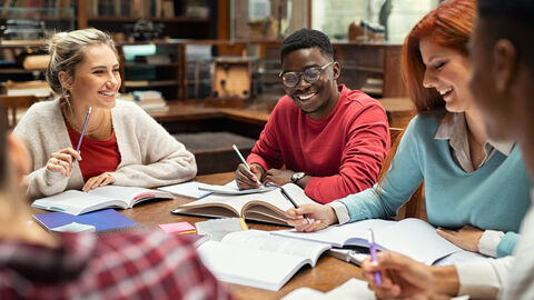 Auf der Abbildung sieht man vier Menschen, die an einem Tisch voller Bücher sitzen und lernen