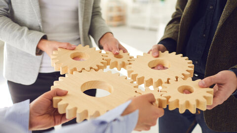 Auf dem Bild sieht man drei Personen, die Zahnräder halten. Die Zahnräder greifen ineinander. Symbolbild Mitwirkungsaufgaben