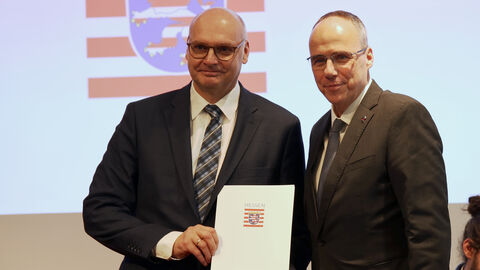 LfV-Präsident Bernd Neumann und der hessische Innenminister Peter Beuth präsentieren Neumanns Ernennungsurkunde. Im Hintergrund ist eine Projektion des Hessenwappens zu sehen.